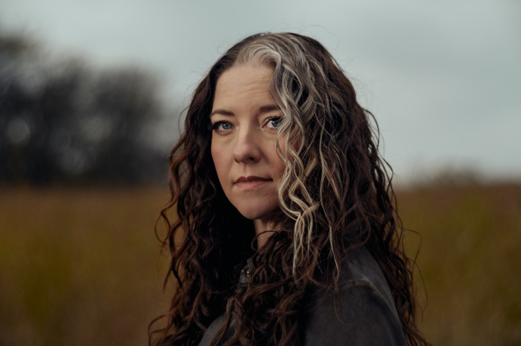 A woman with curly hair and highlights gazes thoughtfully outdoors, with a blurred grassy field and cloudy sky in the background.