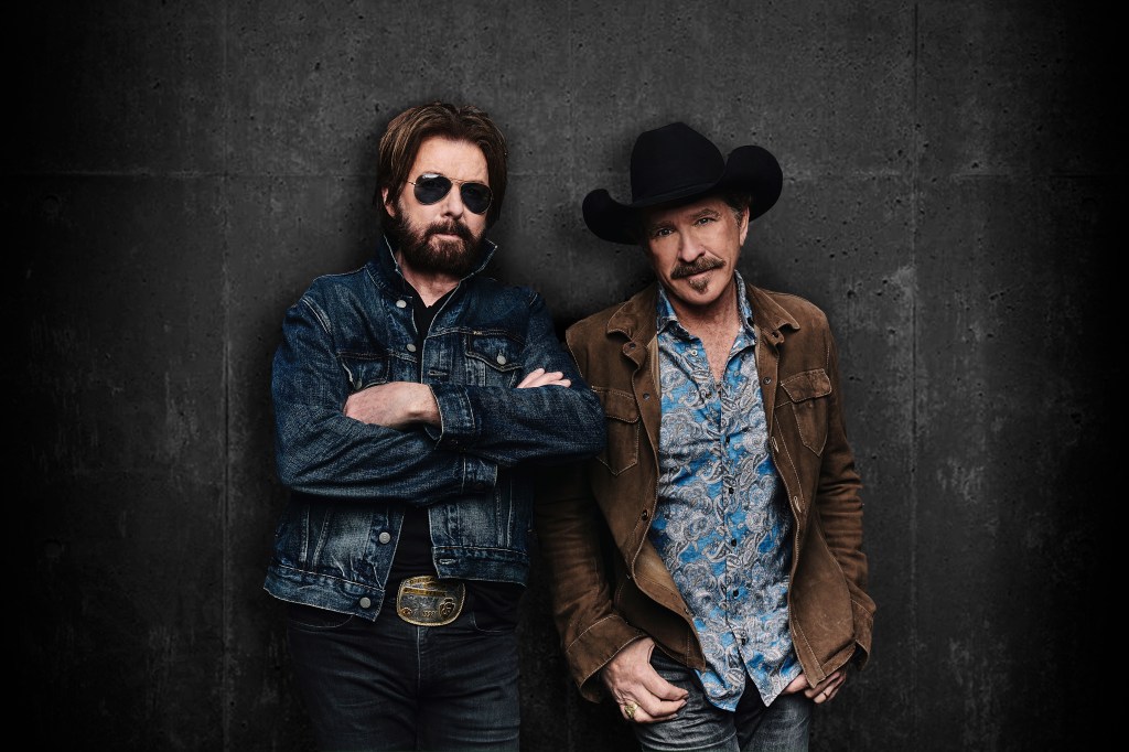 Two men standing side by side against a dark textured background. One man wears a denim jacket and sunglasses, while the other dons a western-style shirt and a cowboy hat.