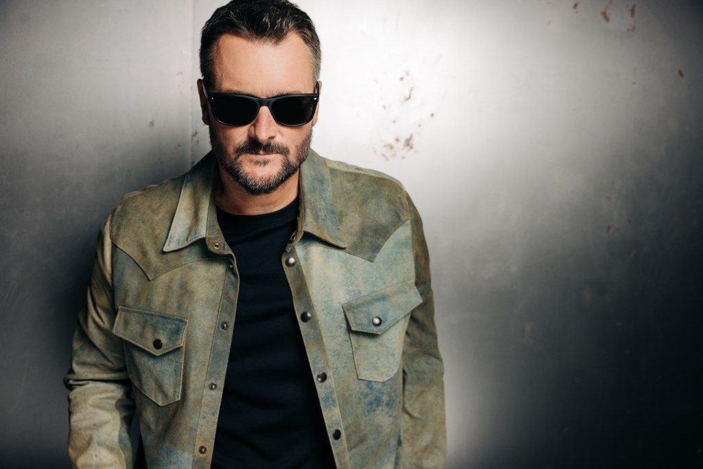 A man with a beard wearing sunglasses and a greenish distressed jacket, posing against a textured gray background.