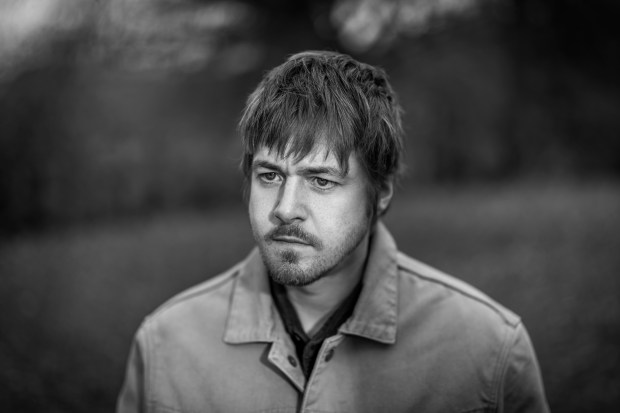 A black and white portrait of a young man looking serious, wearing a light jacket, with a blurred natural background.