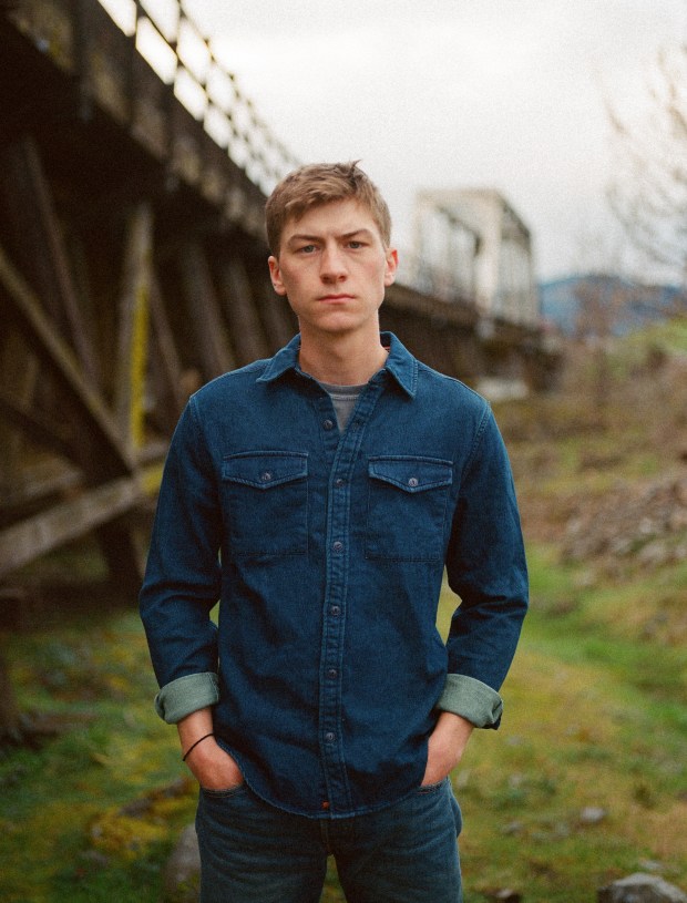 A young man standing outdoors with a serious expression, wearing a blue denim shirt and light-colored pants, in a natural setting near a wooden structure.